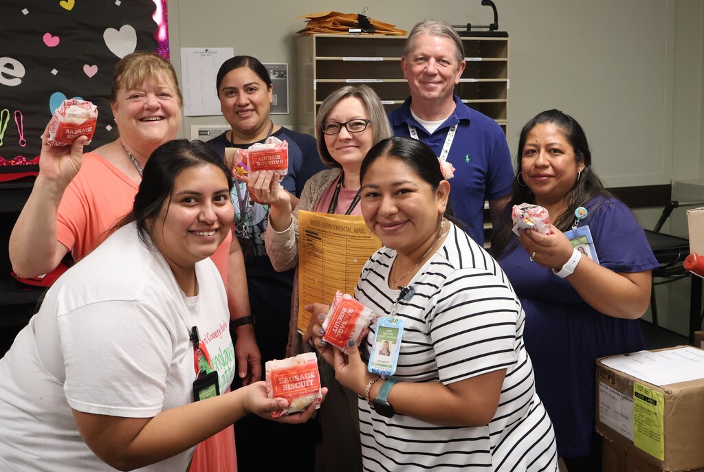 There are seven people who are posed for a picture each holding a sausage biscuit from Hardee's.