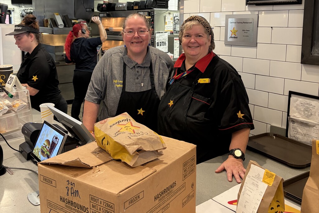 There are two workers at Hardee's standing behind the counter posing for a picture with a box of sausage and biscuits. 