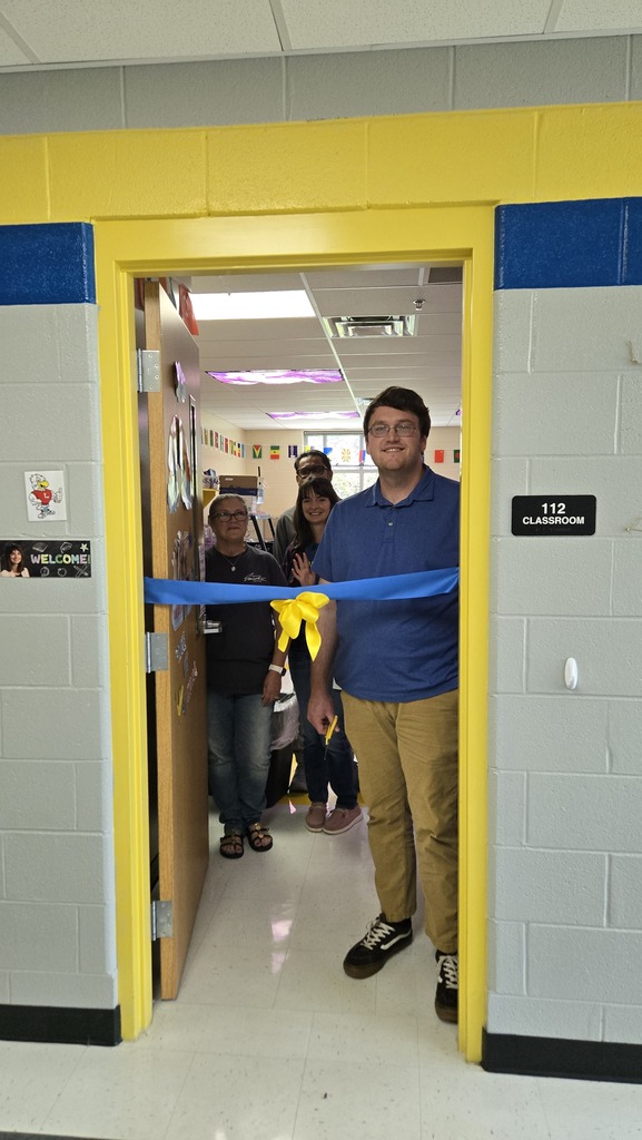 A teacher is standing in the doorway to his classroom with other teachers behind him as he prepares to cut a ceremonial ribbon that is across the doorway. 
