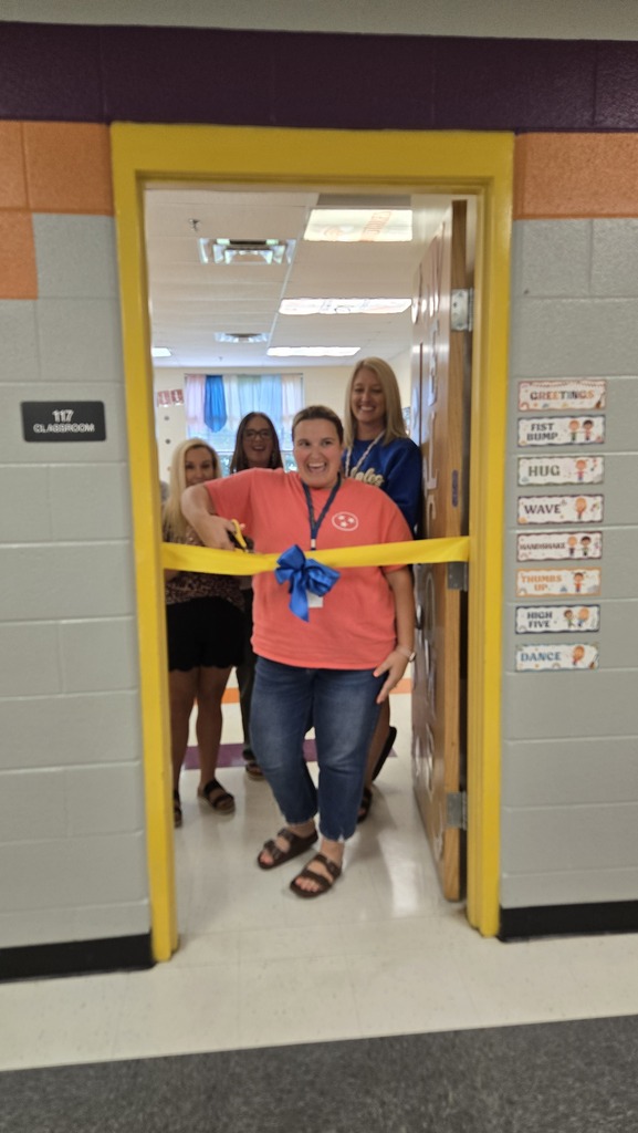 A teacher is standing in the doorway to her classroom with other teachers behind her as she prepares to cut a ceremonial ribbon that is across the doorway. 