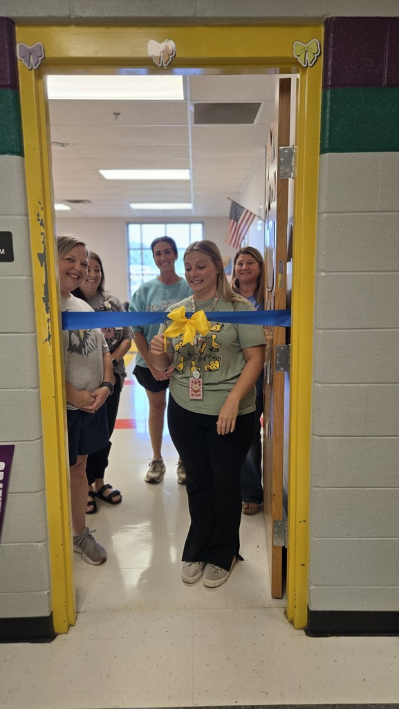 A teacher is standing in the doorway to her classroom with other teachers behind her as she prepares to cut a ceremonial ribbon that is across the doorway. 