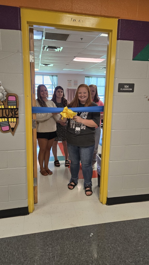 A teacher is standing in the doorway to her classroom with other teachers behind her as she prepares to cut a ceremonial ribbon that is across the doorway. 