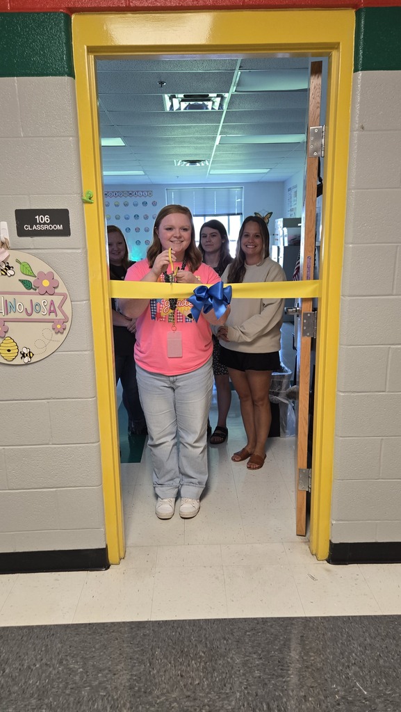 A teacher is standing in the doorway to her classroom with other teachers behind her as she prepares to cut a ceremonial ribbon that is across the doorway. 