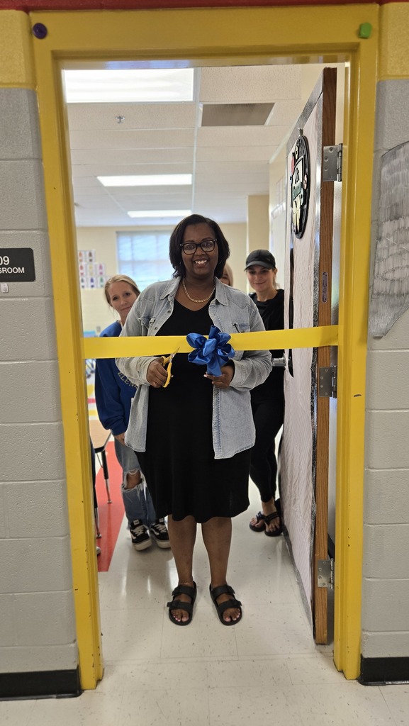 A teacher is standing in the doorway to her classroom with other teachers behind her as she prepares to cut a ceremonial ribbon that is across the doorway. 