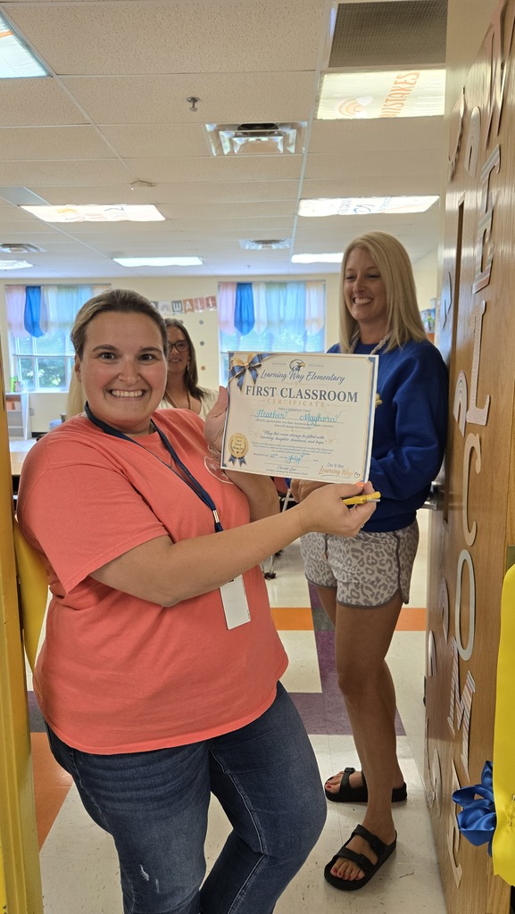 A teacher holds up a certificate about this being her first classroom with a few other teachers standing behind her. 