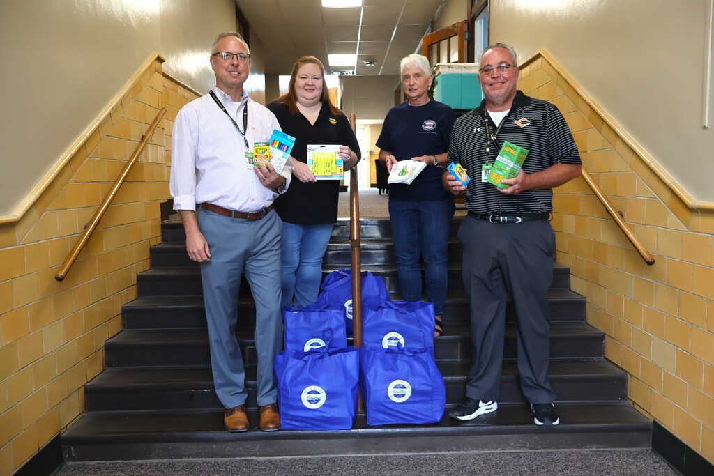 Four people are standing on a set of stairs each holding school supplies while there are five bags placed on the stairs between them. 