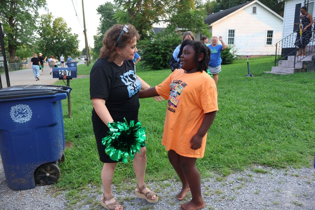 A teacher and a student are standing outside hugging in front of the student's house and the teacher is holding a pom pom in her hand. 