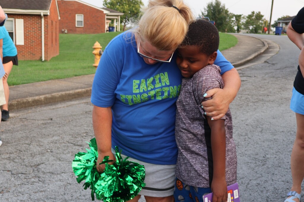 A teacher holding a green pom pom is hugging a student in the street. 