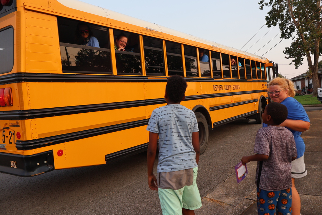 A school bus is parked with the windows down as teachers are looking outside at students who are standing on the side of the road watching with a teacher. 