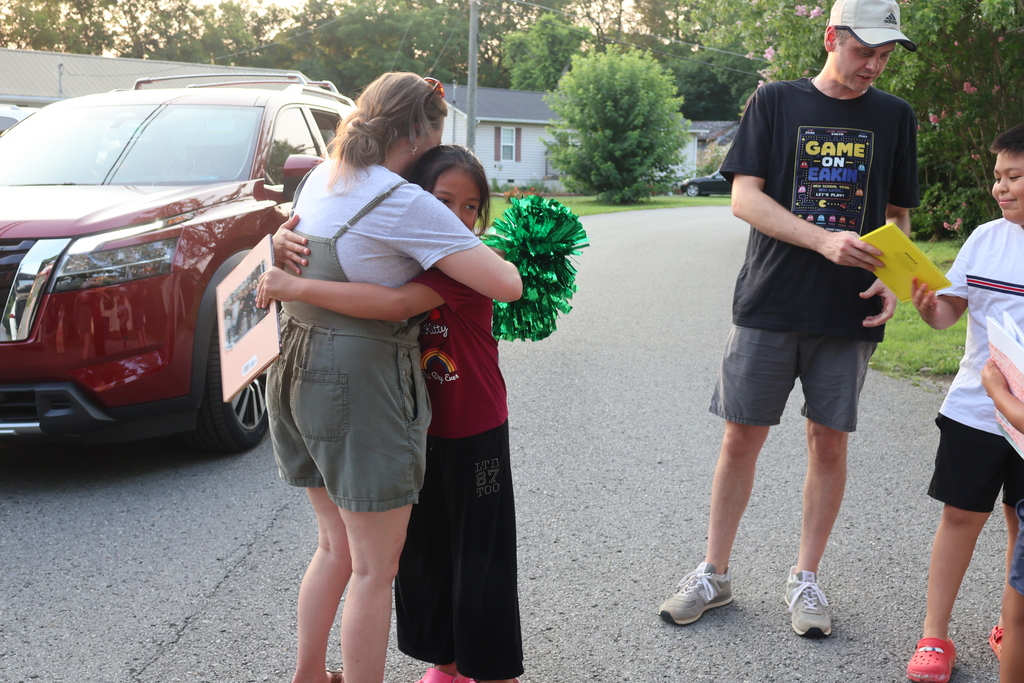 A teacher holding green pom poms is standing in the street hugging a student while another teachers is in the background looking at a book with another student. 