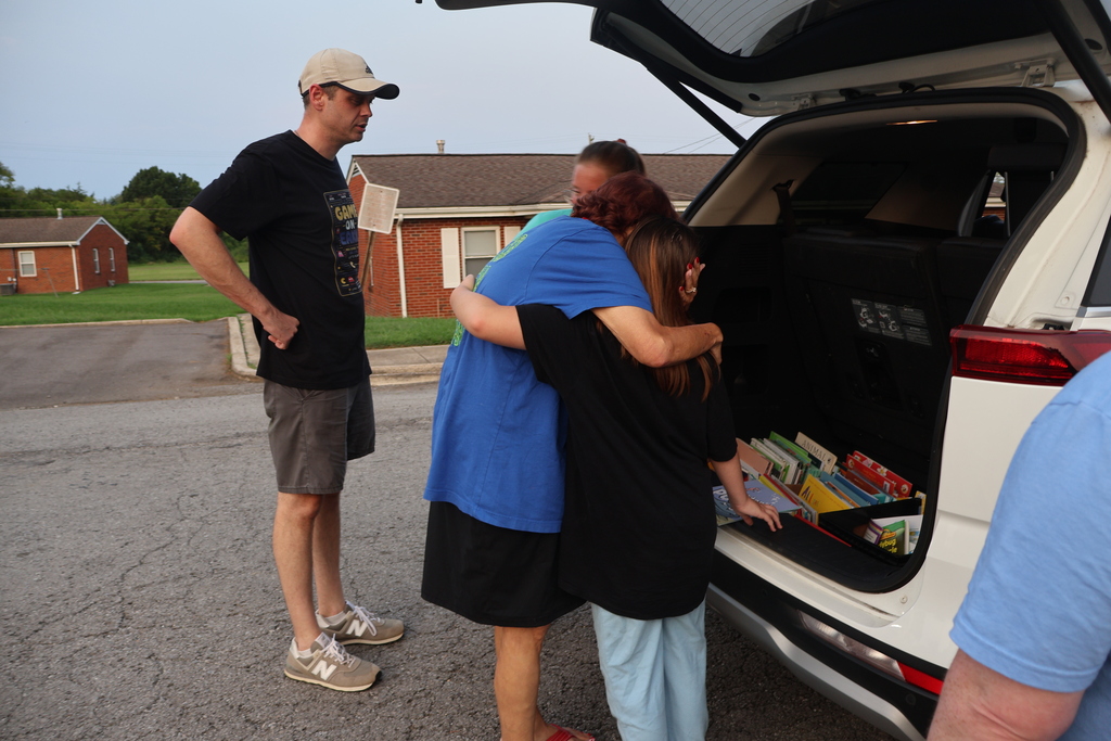 A teacher is hugging a student as they look at books in the back of the van as other teachers are standing nearby. 