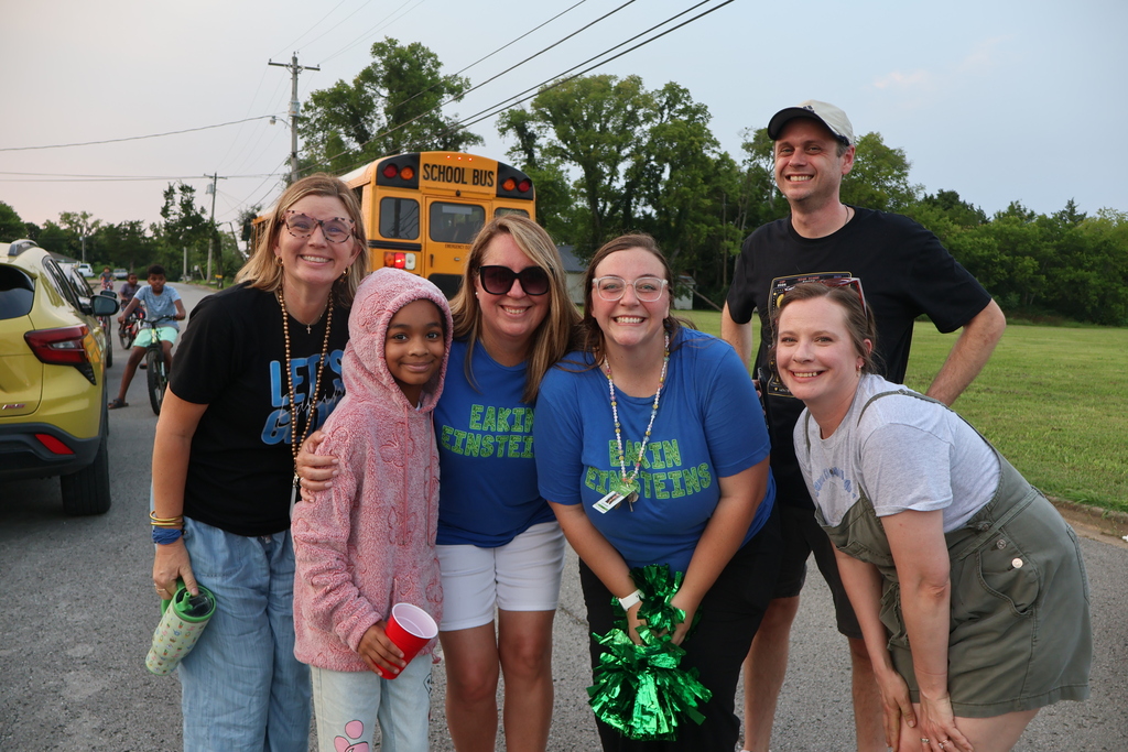 There are five teachers who are all posed for a group picture as they stand in the street with the school bus in the background. 