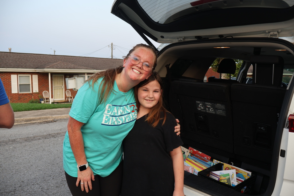 A teacher is posed with a student behind a van that has its trunk open with books in the street. 