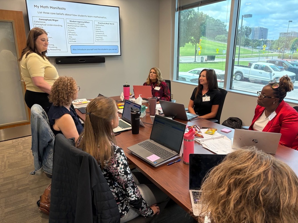 There is a group of around six women who are all seated at a table with their laptops as they listen to another woman presenting to them with tv behind her displaying information. 
