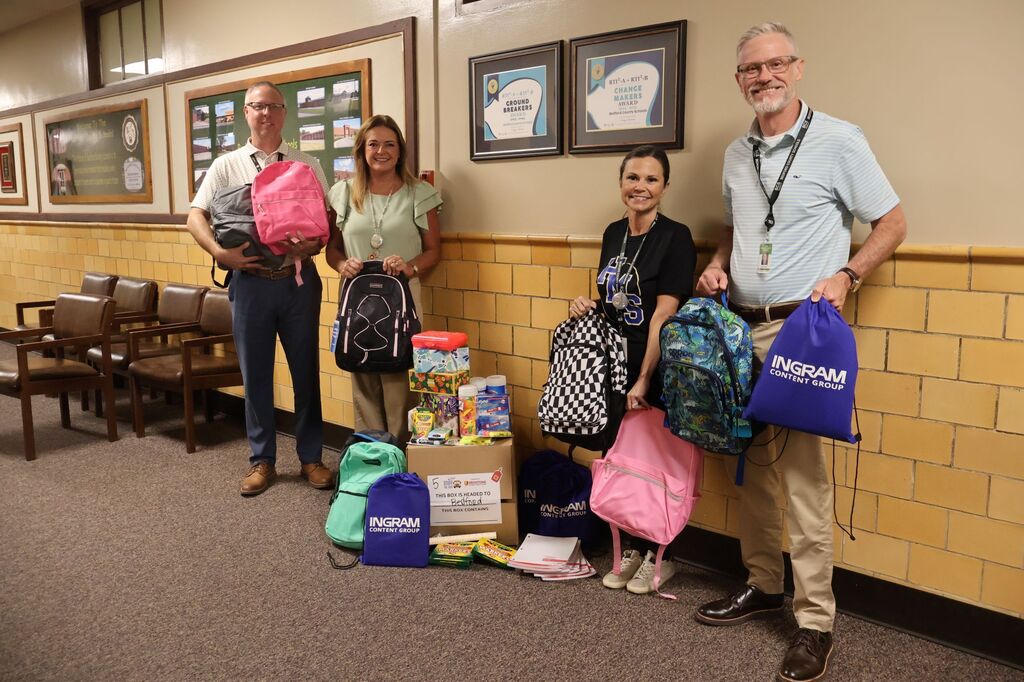 There are four people in this photo posed in the hallway holding up school supplies that were donated. 