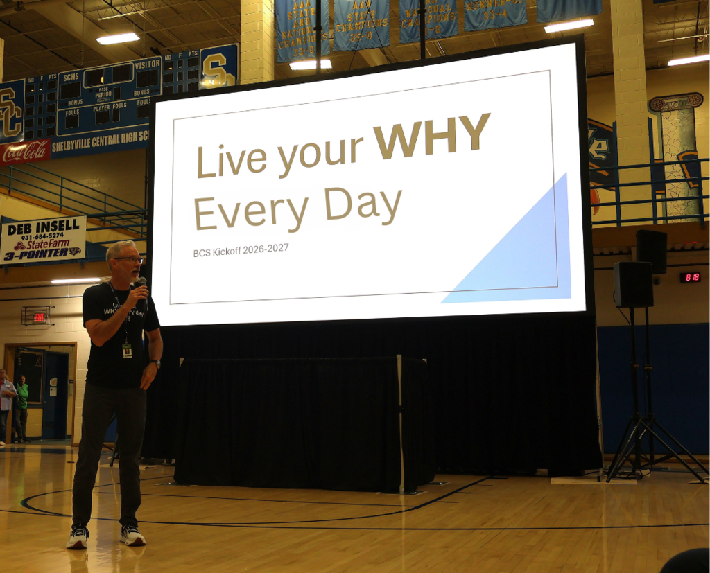 The superintendent is standing near the middle of the gym floor in front of a tall screen that says "Live Your Why Every Day".