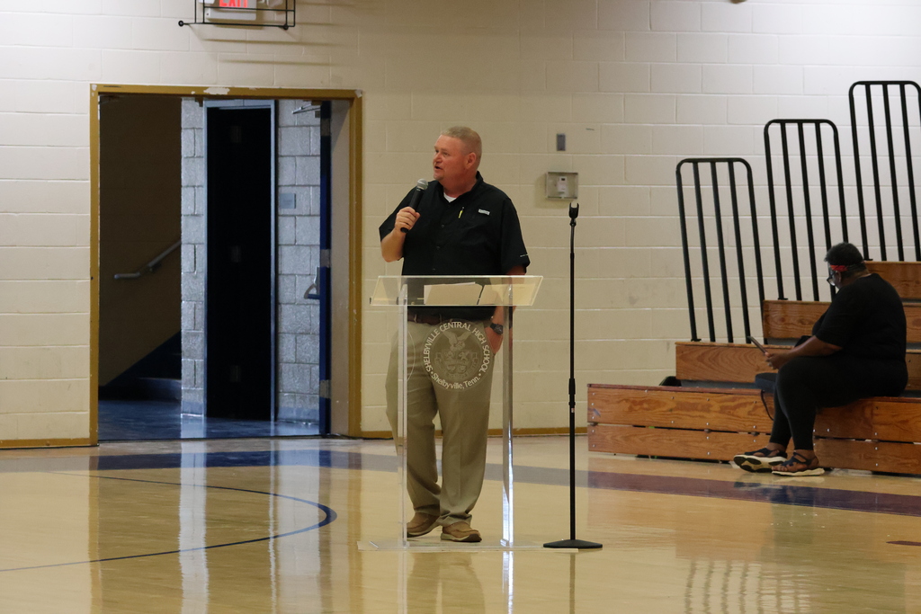 A man is standing on the gym floor at a clear podium holding a microphone in his hand as he speaks to the crowd. 
