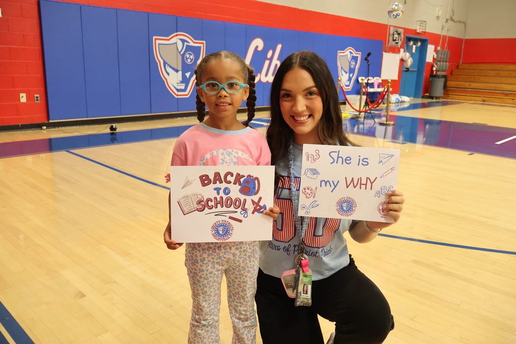 A teacher kneels down beside a student in the gym and they are both holding up signs about back to school. One sign says, "Back to School" and the other says, "She is my WHY".  