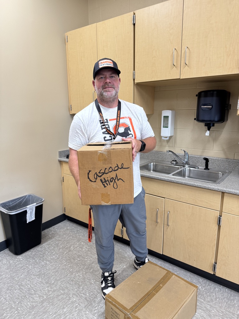 Staff member stands in Cascade High School teacher workroom holding cardboard box labeled "Cascade High" during Musgrave Pencil Company donation dropoff. Another donation box sits on floor in foreground.