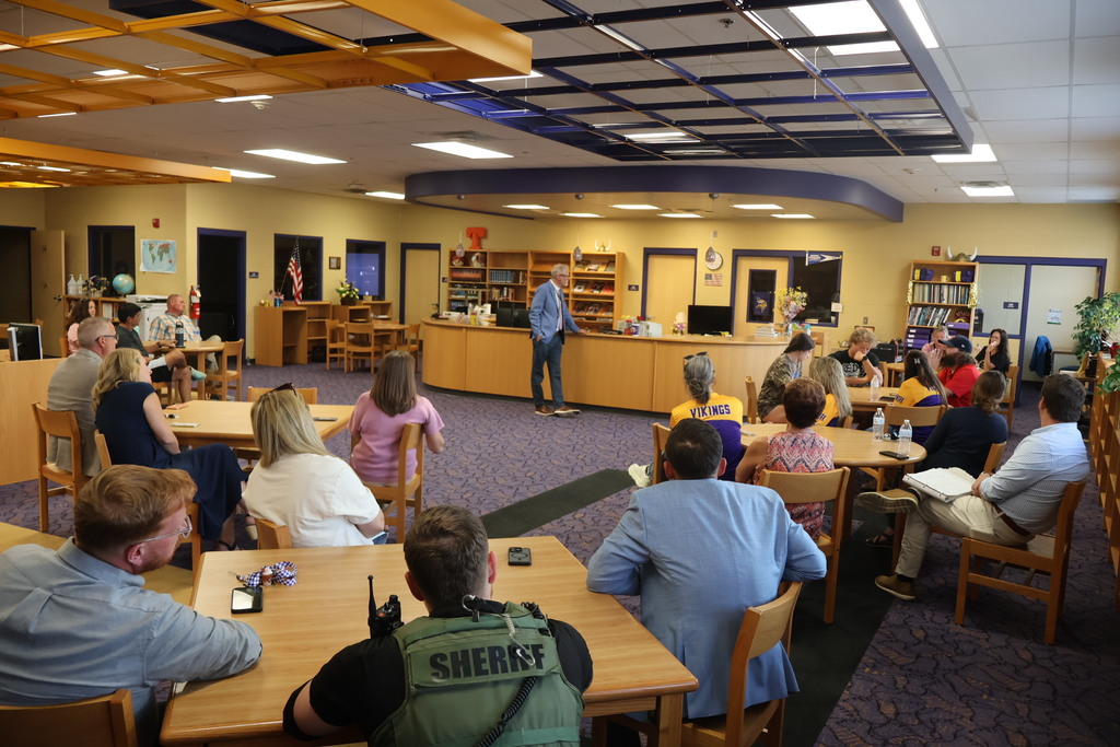 There is a group of people all sitting in the library at tables as the superintendent stands and has a discussion with the individuals. 
