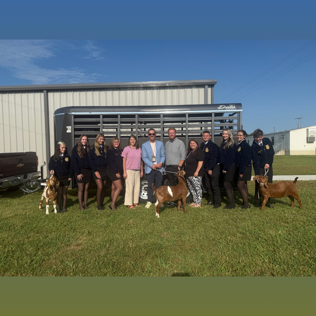 There are several people all posed for a group picture outside in front of a building with everyone posed in front of a livestock trailer with a few goats that they are holding.