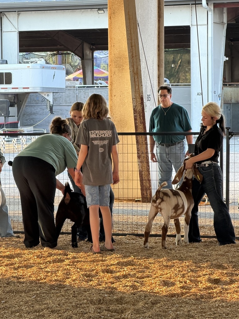 There are two females in this picture who are each holding a goat at a goat show as other are looking on.