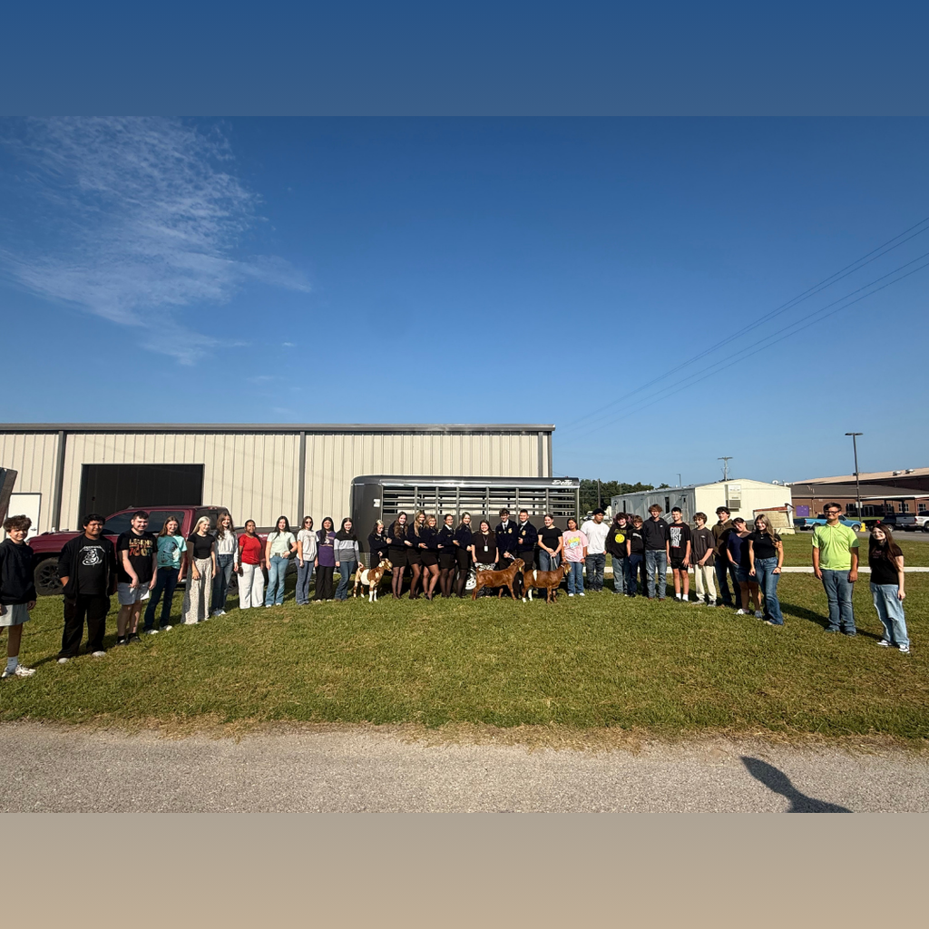This is a large group picture outside in front of a building with everyone posed in front of a livestock trailer with a few goats that they are holding.