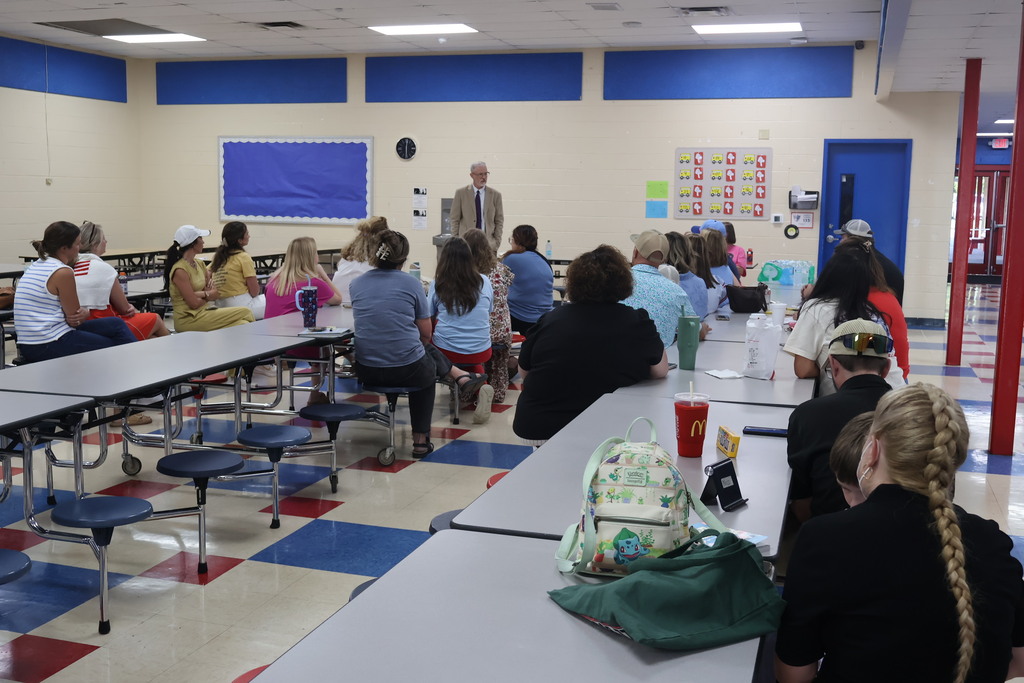 This is a picture of several people all sitting down at tables in the lunchroom as the superintendent speaks to them. 