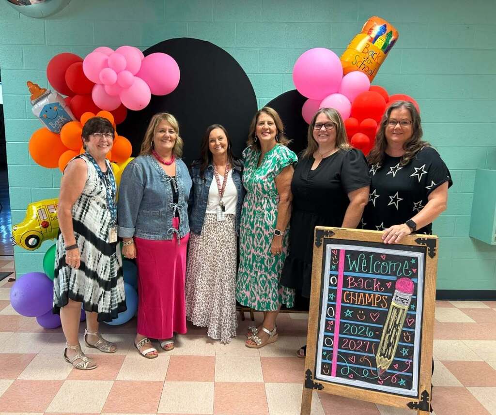 There are six people in this picture who are all posed for a group picture in the hallway in front of a balloon arch and small folding sign.