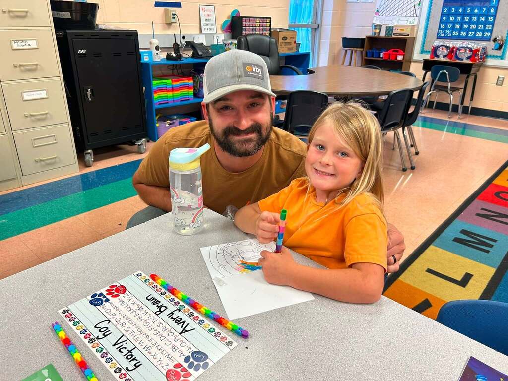 There is an adult and a child who are sitting in chairs at a table in a classroom.