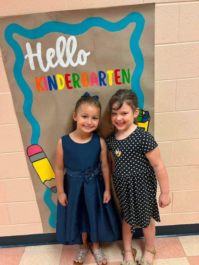 There are two little girls who are posed for a picture in the hallway by a sign that says "Hello Kindergarten".