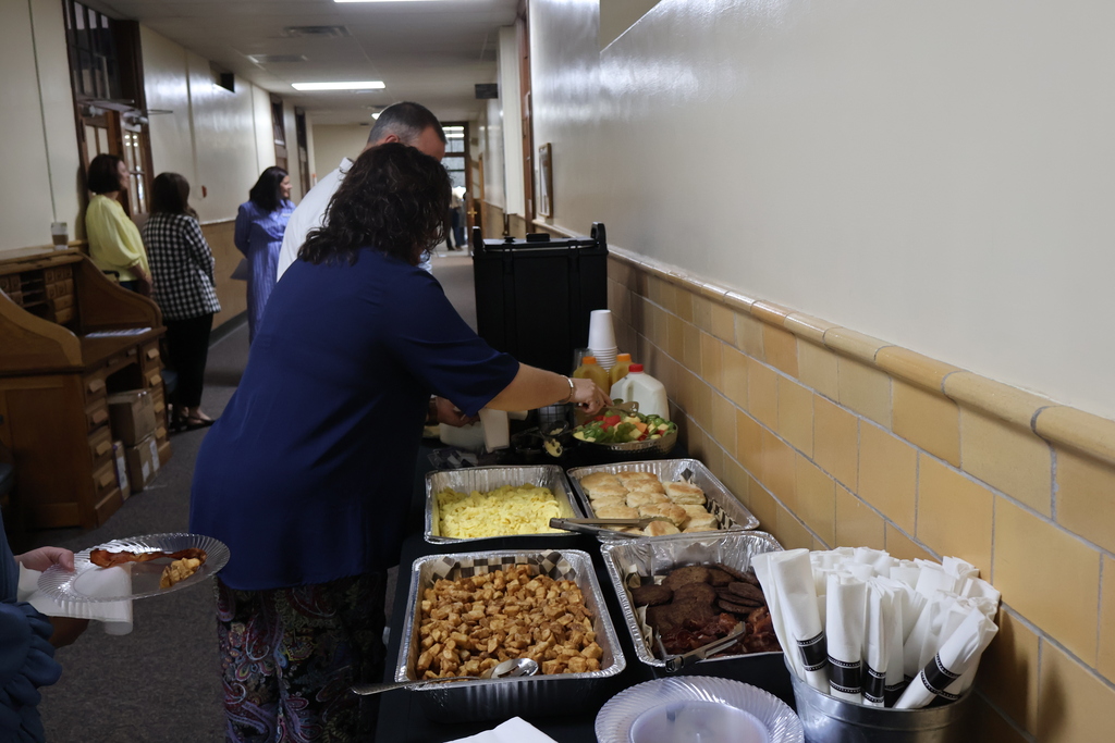 There are several people in the hallway of a building where there is a table that has food on it.