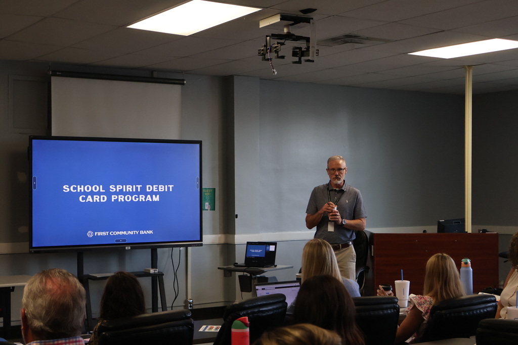 This is a picture of the superintendent who is speaking to the principals with a large screen behind him that says "School Spirit Debit Card Program". 