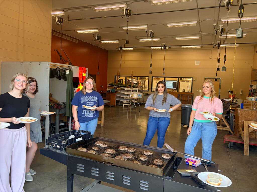 Veterinary science students holding plates of food next to large industrial griddle cooking hamburgers after hands-on class activity.