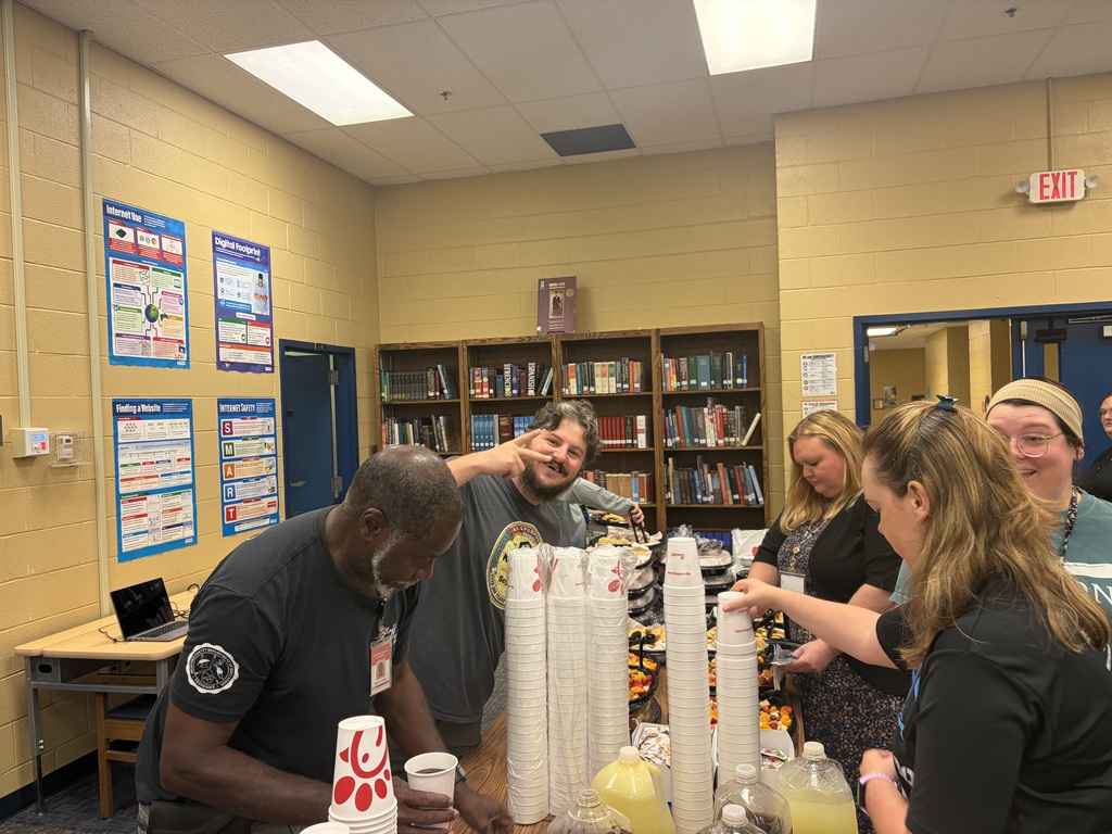 There are several people standing in the library around a table that has food on it from Chic-fil-A.