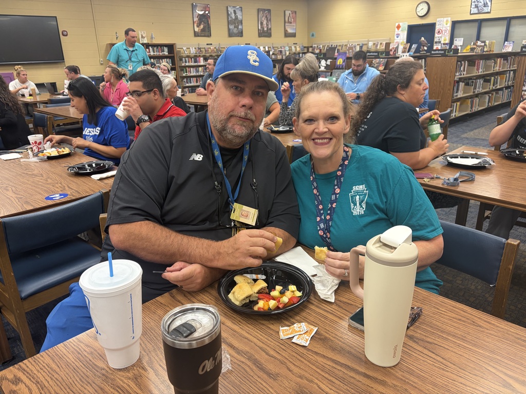This is a picture of two people who are sitting at a table in the library eating breakfast while other people are also sitting at tables in the background.