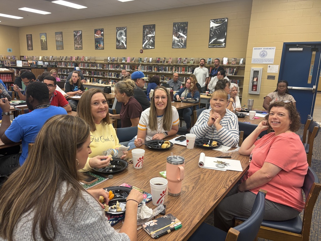 This is a picture of five people who are sitting at a table in the library eating breakfast while other people are also sitting at tables in the background.