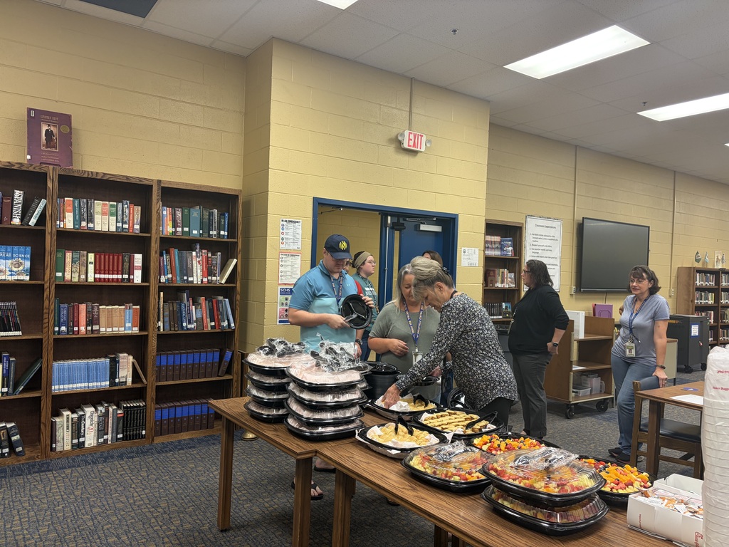 There are several people standing in the library around a table that has food on it from Chic-fil-A.