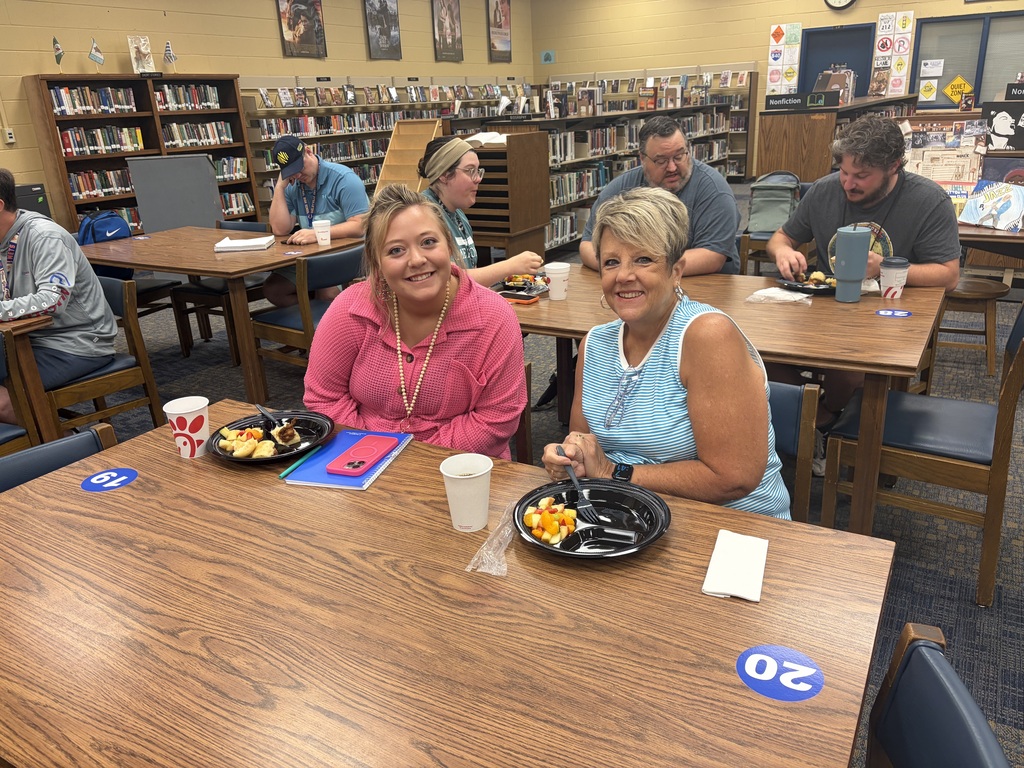 This is a picture of two people who are sitting at a table in the library eating breakfast while other people are also sitting at tables in the background.