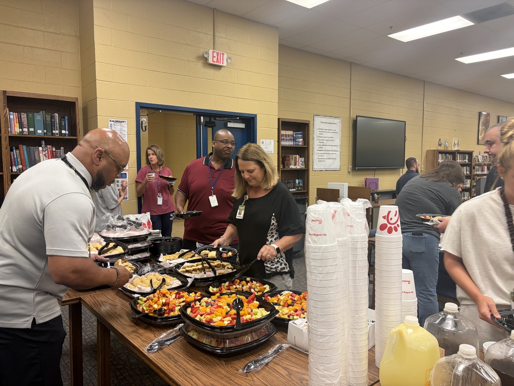 There are several people standing in the library around a table that has food on it from Chic-fil-A.