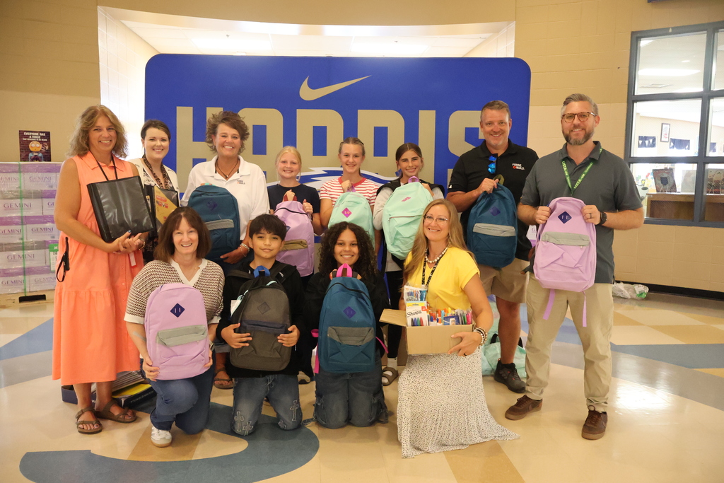 This is a group picture where everyone is either holding a backpack or school supplies that were donated by Forge & Build. They are standing in the hallway in front of a large banner that says "Harris Eagles". 