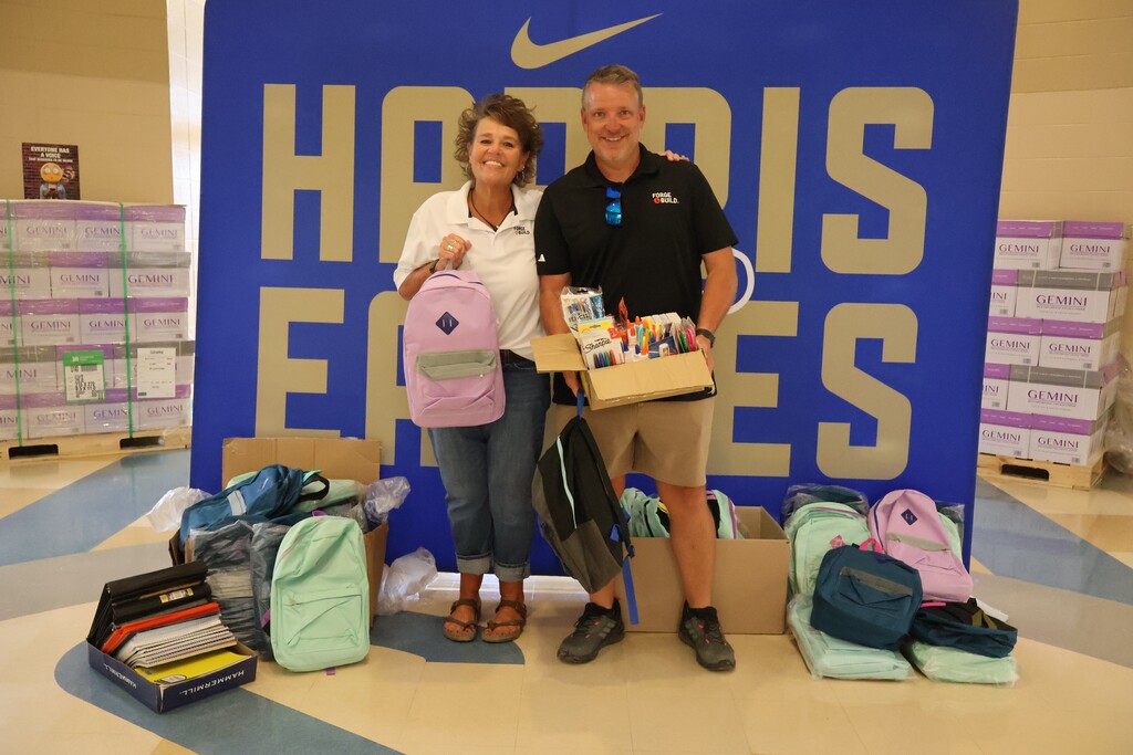 There are two people posed for a picture in the hallway in front of the large banner that says "Harris Eagles" and they are each holding backpacks while one is also holding a box of school supplies. Backpacks are on the ground around them. 