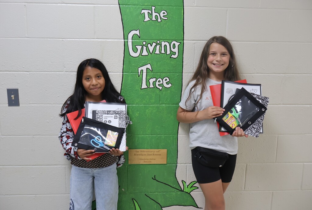This is a picture of two students who are posed for a group picture in the hallway of their school and each are holding up school supplies they received. 