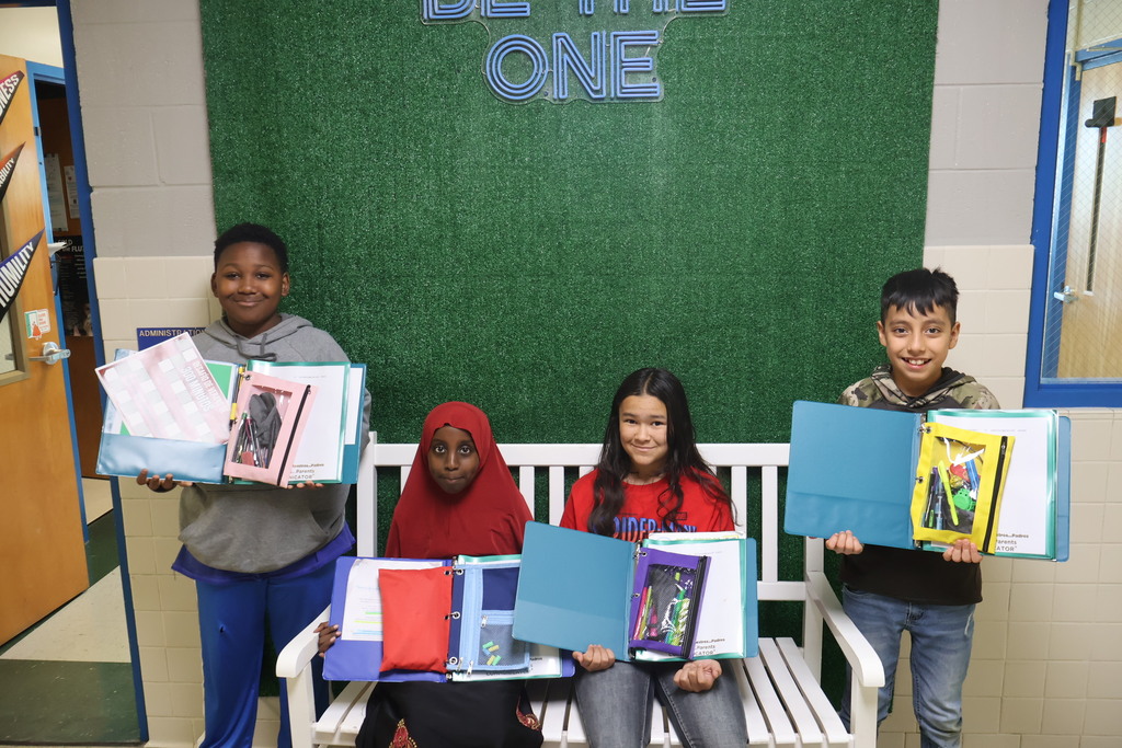 This is a picture of four students who are all posed for a group picture in the hallway of their school and each are holding up school supplies they received.
