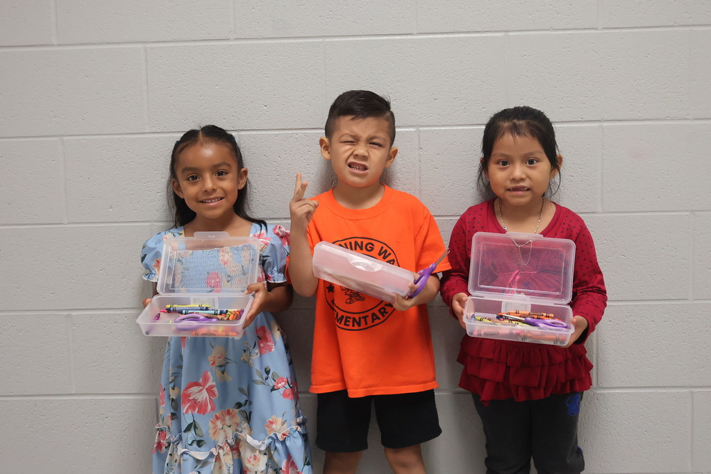 This is a picture of three students who are all posed for a group picture in the hallway of their school and each are holding up school supplies they received.