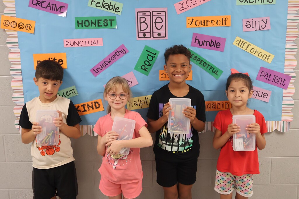 This is a picture of four students who are all posed for a group picture in the hallway of their school and each are holding up school supplies they received.