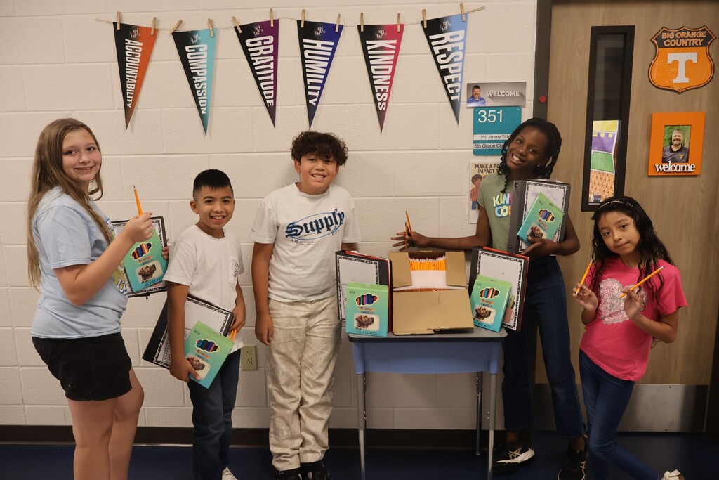 This is a picture of five students who are all posed for a group picture in the hallway of their school and each are holding up school supplies they received. 