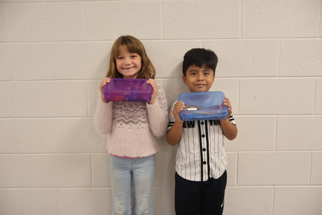 This is a picture of two students who are posed for a group picture in the hallway of their school and each are holding up school supplies they received. 
