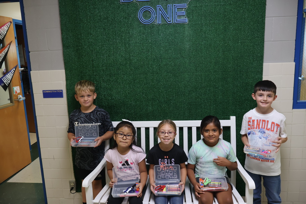 This is a picture of four students who are all posed for a group picture in the hallway of their school and each are holding up school supplies they received. 