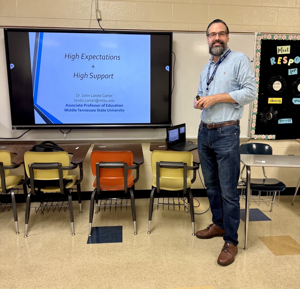 A man is standing in a classroom by a digital board that has a PowerPoint slideshow on behind him.